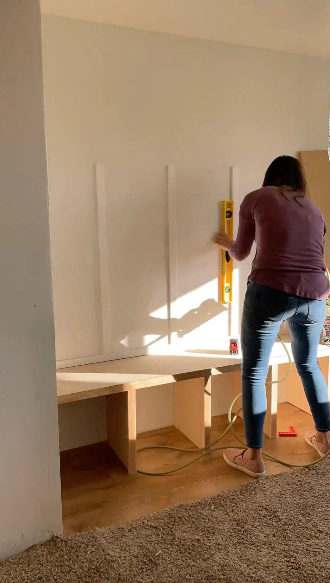 A woman installing vertical wooden slats on a wall above a built-in storage bench, using a level to ensure precision. The DIY project is coming together with clean lines, adding a custom, modern touch to the space.